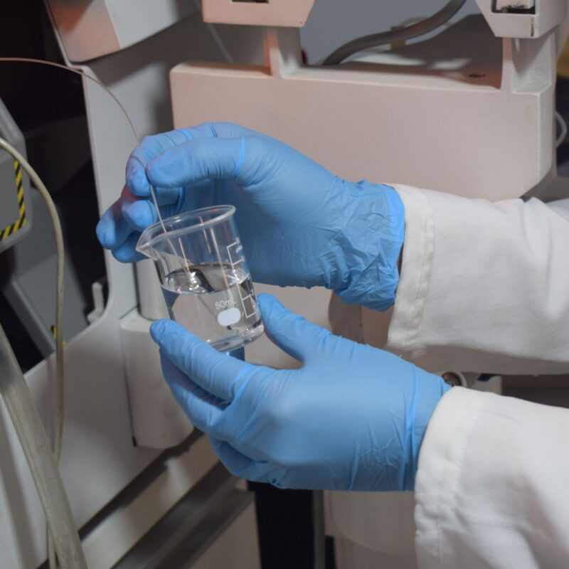 Gloved hands test liquids in a beaker in a lab.